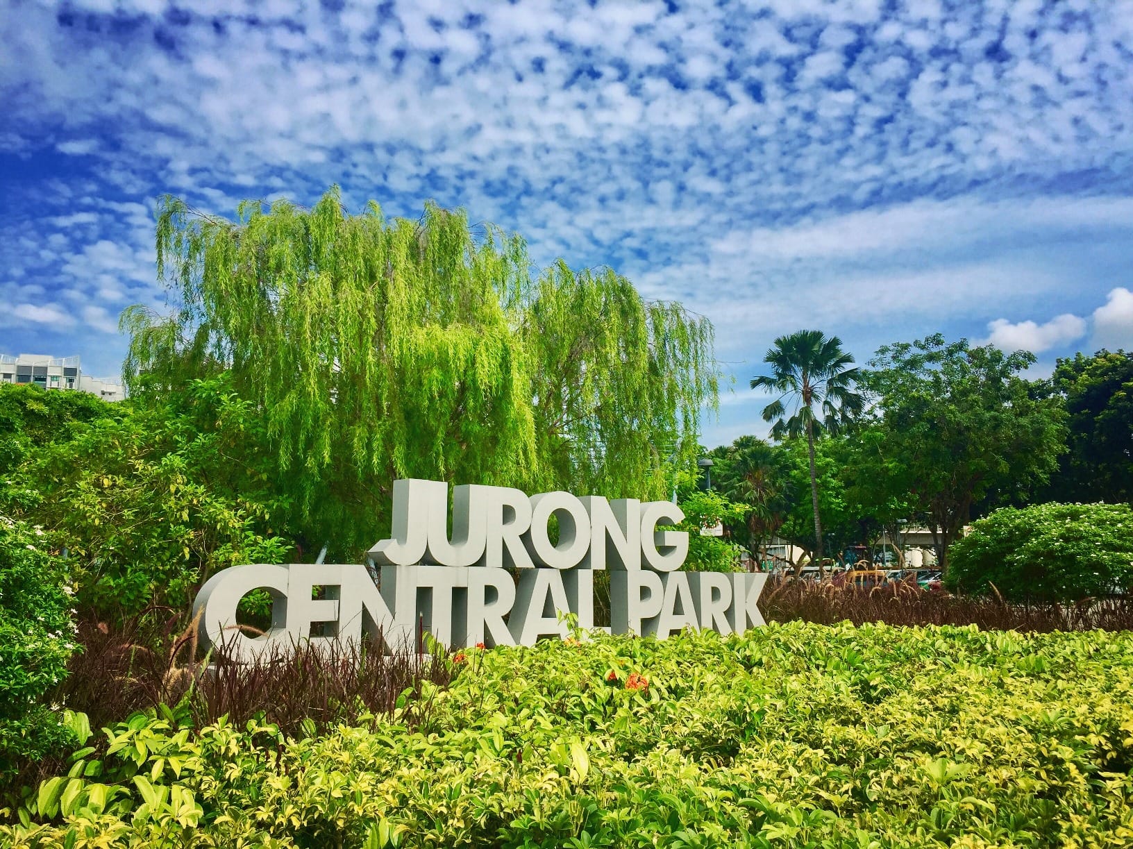 Large white JURONG CENTRAL PARK sign amid green shrubs and trees under a blue, cloud-filled sky